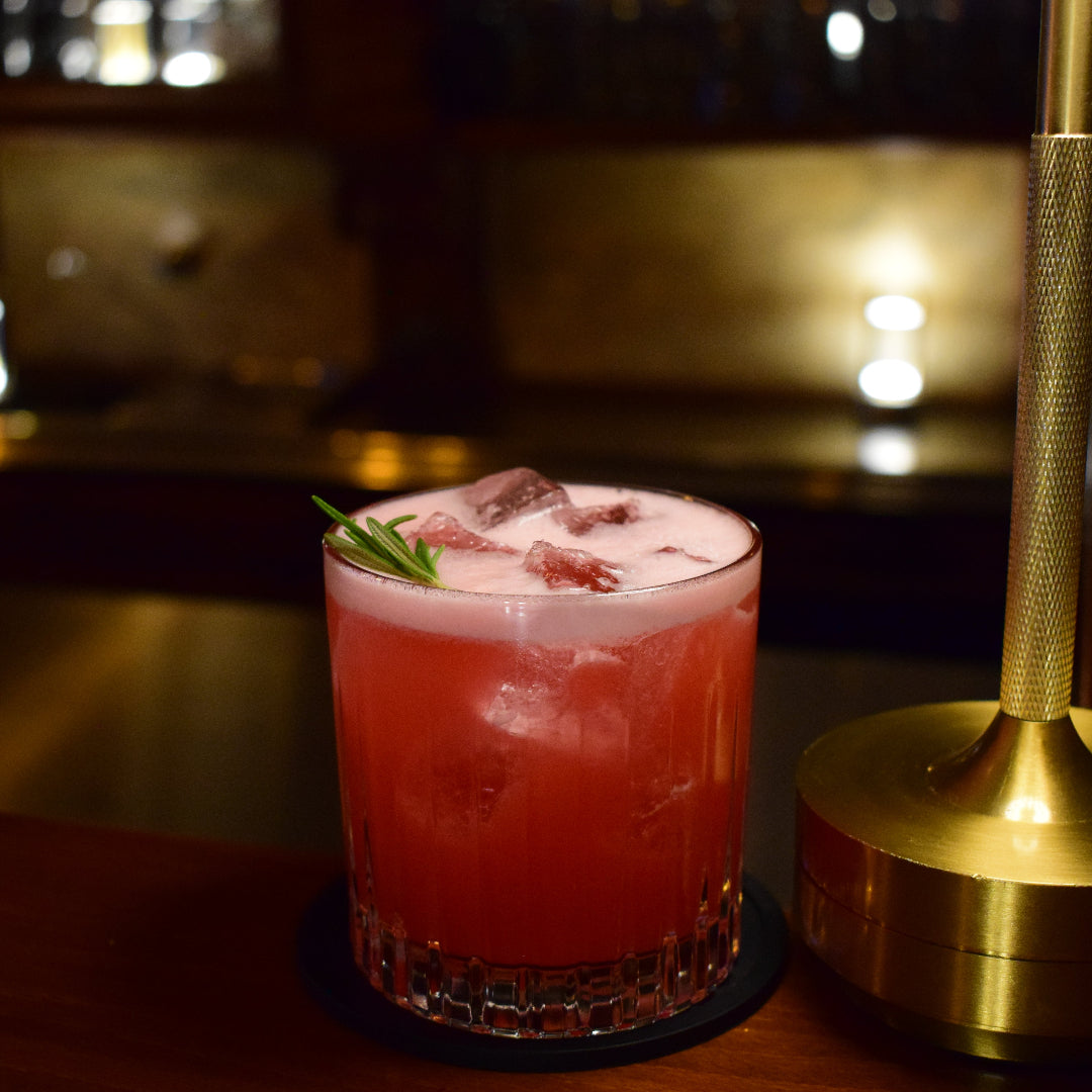 Pink cocktail with ice and a sprig of rosemary on a bar counter with a blurred background