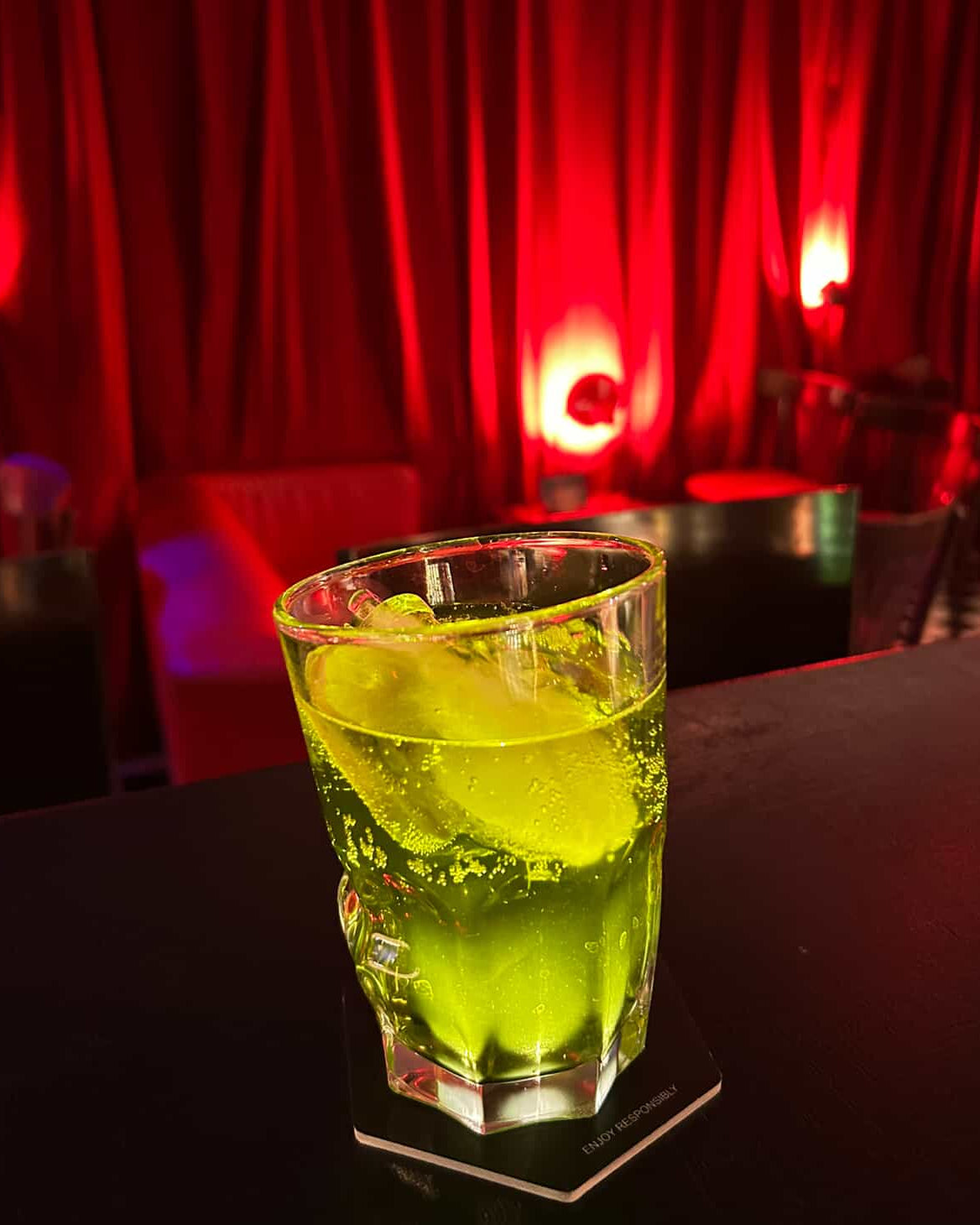 Green cocktail with ice in a glass on a coaster, set against a red curtain background.
